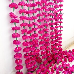 May include: Rows of vibrant pink artificial marigold flower garlands. Each flower is attached to a green stem, creating a dense, colorful display. The garlands are hanging against a white wall, with some lying on the floor. The text "Craft colours India" is visible.