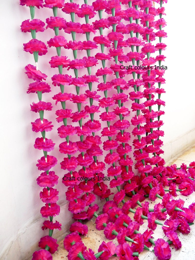 May include: Rows of vibrant pink artificial marigold flower garlands. Each flower is attached to a green stem, creating a dense, colorful display. The garlands are hanging against a white wall, with some lying on the floor. The text "Craft colours India" is visible.