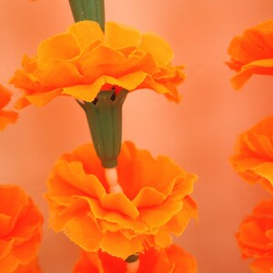 Puede incluir: Primer plano de una guirnalda de flores de cal&eacute;ndula de color naranja brillante. Las flores tienen m&uacute;ltiples capas de p&eacute;talos y est&aacute;n unidas a tallos verdes. El fondo es de color naranja suave, creando una sensaci&oacute;n c&aacute;lida y acogedora.