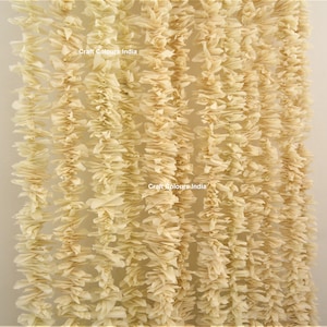 May include: A close-up of a cream-colored garland made of paper. The garland is made of many small, ruffled pieces of paper that are strung together. The garland is hanging from a white wall. The text "Craft Colours India" is visible on the garland.