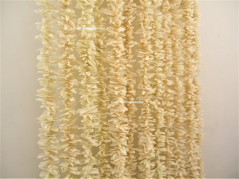 May include: A close-up of a cream-colored garland made of paper. The garland is made of many small, ruffled pieces of paper that are strung together. The garland is hanging from a white wall. The text "Craft Colours India" is visible on the garland.