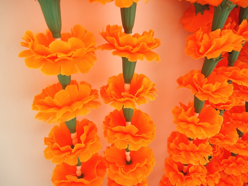 Puede incluir: Primer plano de guirnaldas de flores de cal&eacute;ndula de color naranja vibrante. Cada guirnalda presenta m&uacute;ltiples flores de color naranja brillante con tallos verdes. Las guirnaldas probablemente se utilizan para la decoraci&oacute;n o ceremonias religiosas.