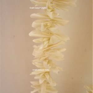 May include: A long garland of cream-colored paper flowers, hanging from the ceiling. The garland is made of many individual paper flowers, each with a slightly different shape. The garland is decorated with the text "Craft Colours India" twice.