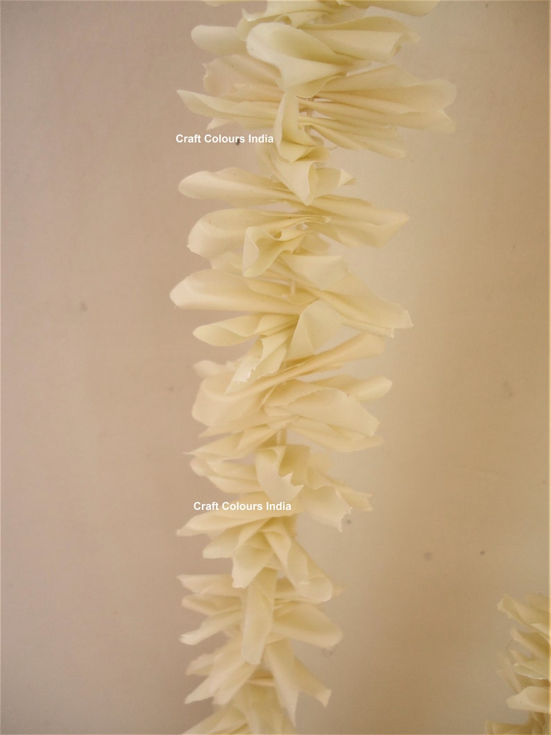 May include: A long garland of cream-colored paper flowers, hanging from the ceiling. The garland is made of many individual paper flowers, each with a slightly different shape. The garland is decorated with the text "Craft Colours India" twice.
