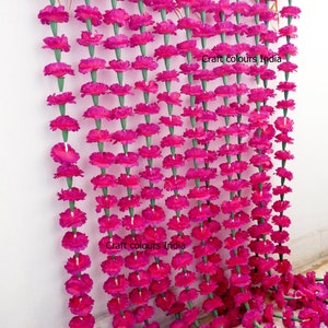 May include: Rows of vibrant fuchsia marigold flower garlands hang against a white wall. The garlands are made of many individual flowers with green stems. The text "Craft colours India" is visible on the wall.