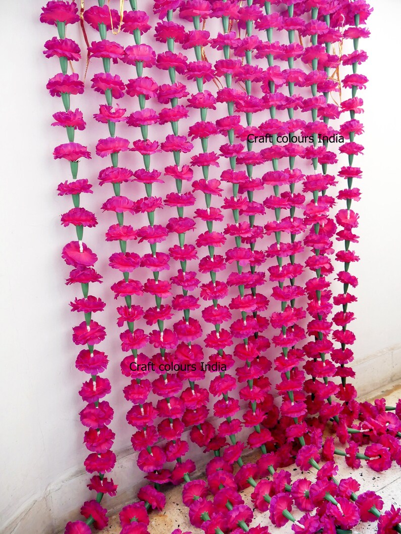 May include: Rows of vibrant fuchsia marigold flower garlands hang against a white wall. The garlands are made of many individual flowers with green stems. The text "Craft colours India" is visible on the wall.