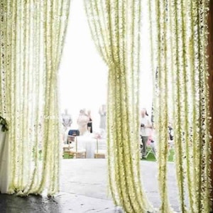 May include: White flower garlands hang from the ceiling, creating a curtain-like effect. The garlands are made of artificial flowers and are used as a decorative element for a wedding or other special event.