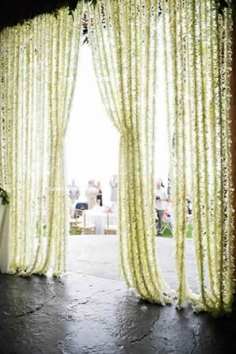 May include: White flower garlands hang from the ceiling, creating a curtain-like effect. The garlands are made of artificial flowers and are used as a decorative element for a wedding or other special event.