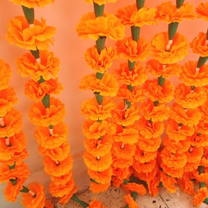 Puede incluir: Primer plano de hileras de guirnaldas de cal&eacute;ndulas de color naranja brillante. Cada flor est&aacute; cuidadosamente dispuesta, creando una exhibici&oacute;n densa y texturizada. Los tallos verdes y los acentos blancos contrastan con las flores naranjas.
