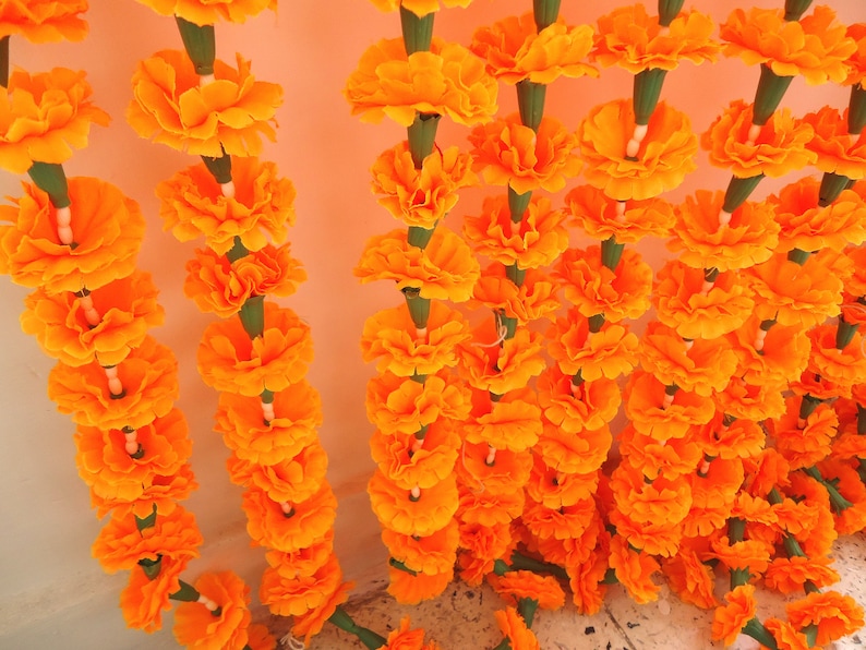 Puede incluir: Primer plano de hileras de guirnaldas de cal&eacute;ndulas de color naranja brillante. Cada flor est&aacute; cuidadosamente dispuesta, creando una exhibici&oacute;n densa y texturizada. Los tallos verdes y los acentos blancos contrastan con las flores naranjas.