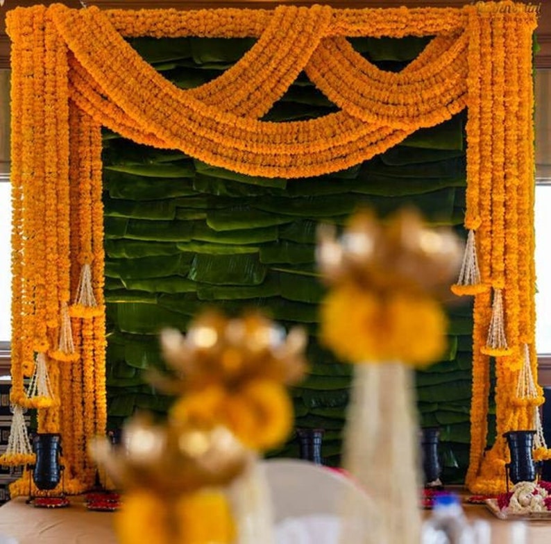 May include: A green backdrop decorated with yellow marigold flower garlands, used for a wedding ceremony. The garlands are draped in a decorative pattern and hang from the ceiling.