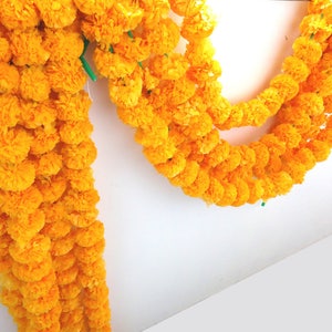 May include: A garland of bright yellow marigold flowers strung together with green string. The garland is hanging on a white wall.