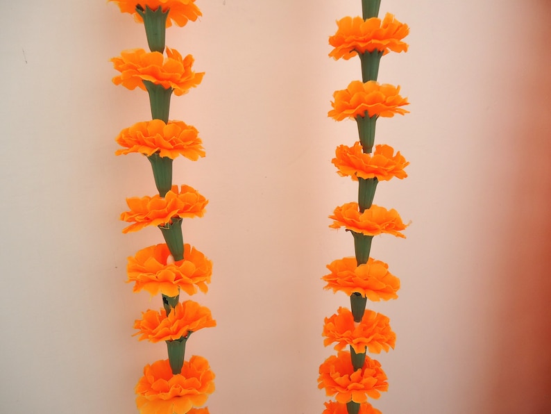 Puede incluir: Dos guirnaldas verticales de cal&eacute;ndulas de color naranja brillante, cada una con m&uacute;ltiples flores y tallos verdes, sobre un fondo claro. Las flores completamente florecidas crean una exhibici&oacute;n vibrante y festiva. Decoraci&oacute;n tradicional.