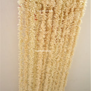 May include: A white paper garland with a ruffled texture, hanging from a wooden dowel. The garland is made up of multiple strands, each with a delicate, frilly design. The text "Craft Colours India" is visible on the garland.