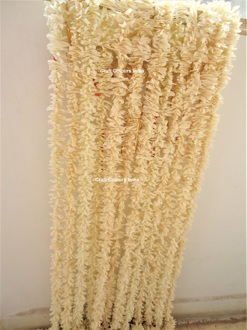 May include: A white paper garland with a ruffled texture, hanging from a wooden dowel. The garland is made up of multiple strands, each with a delicate, frilly design. The text "Craft Colours India" is visible on the garland.