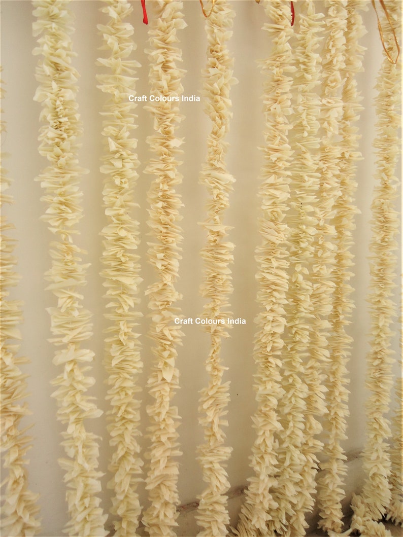 May include: A close-up of a wall decorated with multiple strands of cream-colored paper flower garlands. The garlands are hanging from the ceiling and are arranged in a vertical pattern. The text "Craft Colours India" is visible twice on the wall.