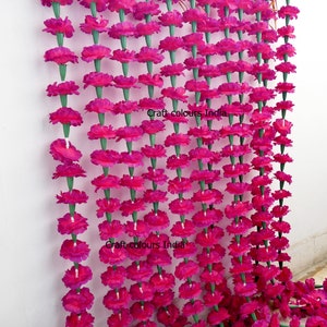 May include: Rows of vibrant fuchsia artificial marigold flower garlands. Each flower is strung together with green stems, creating a dense, colorful display. The garlands are hanging against a white wall, perfect for festive decorations.