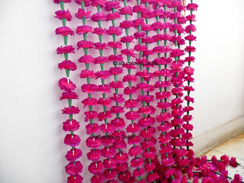 May include: Rows of vibrant fuchsia artificial marigold flower garlands. Each flower is strung together with green stems, creating a dense, colorful display. The garlands are hanging against a white wall, perfect for festive decorations.