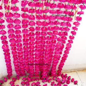 May include: Rows of vibrant fuchsia artificial flower garlands hang from the top of the frame, cascading downwards. The garlands are made of many individual flowers with green stems. The text "Craft colours India" is visible on the image.