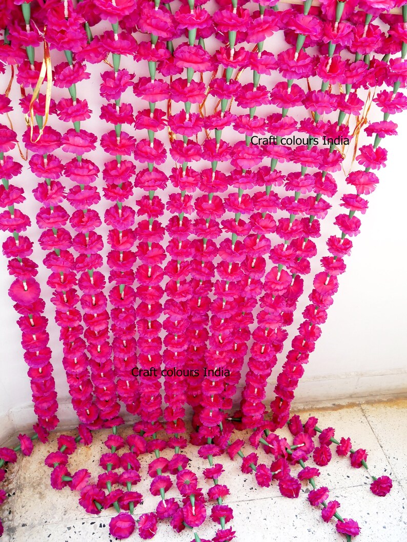 May include: Rows of vibrant fuchsia artificial flower garlands hang from the top of the frame, cascading downwards. The garlands are made of many individual flowers with green stems. The text "Craft colours India" is visible on the image.