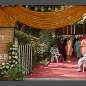 May include: A wooden sign with the text "Preeti Weds Jatinder" decorated with yellow and white flowers. The sign is hanging on a wall with a garland of yellow flowers above it.