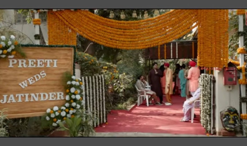 May include: A wooden sign with the text "Preeti Weds Jatinder" decorated with yellow and white flowers. The sign is hanging on a wall with a garland of yellow flowers above it.