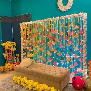 May include: A decorative scene with a beige tufted bench and a garland of yellow roses. The backdrop features hanging decorations in pink, orange, and green, with fairy lights. The teal wall has a white wreath hanging above.