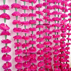 May include: Rows of vibrant pink artificial marigold flower garlands. The flowers are densely packed, with green stems. The garlands are hanging against a white background. The text "Craft colours India" is visible on the image.