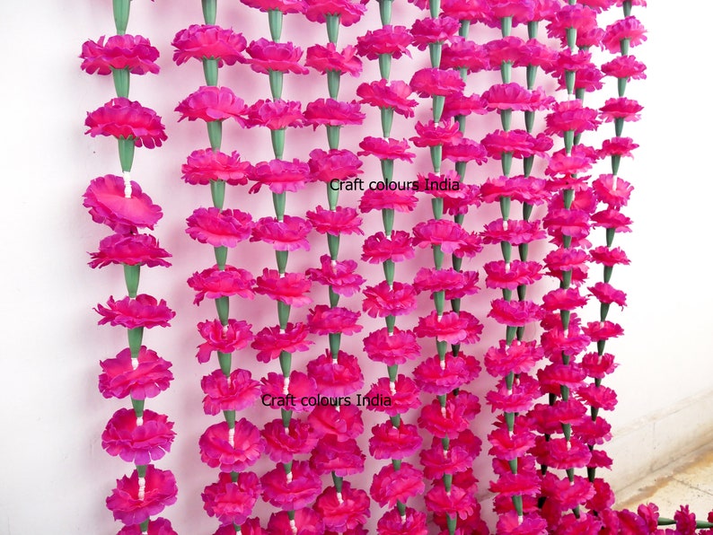 May include: Rows of vibrant pink artificial marigold flower garlands. The flowers are densely packed, with green stems. The garlands are hanging against a white background. The text "Craft colours India" is visible on the image.