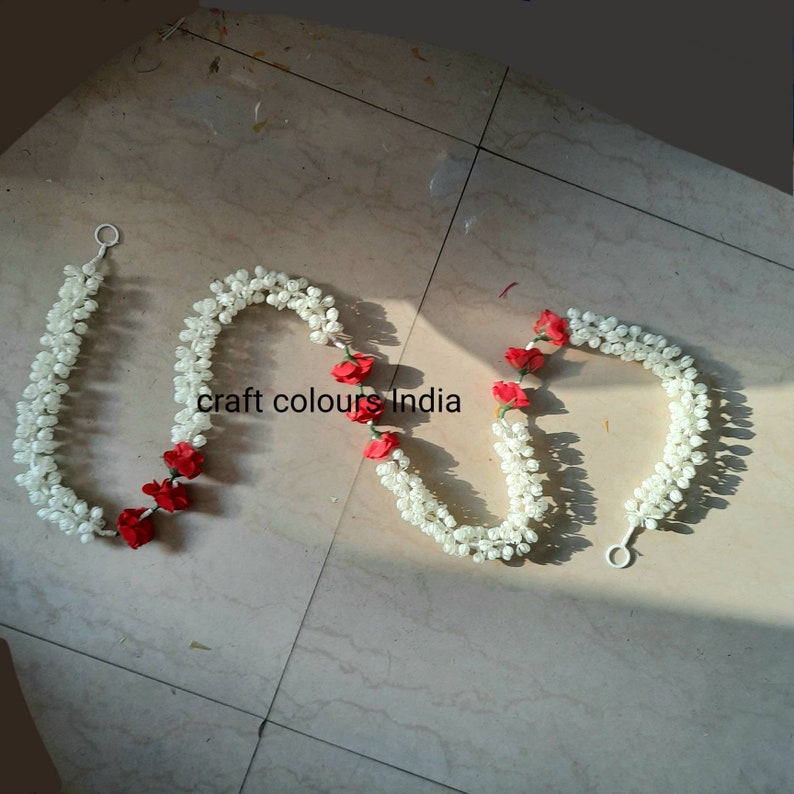 May include: Two white flower garlands with red rose accents. The garlands are made of artificial flowers and are strung together with a loop at each end.