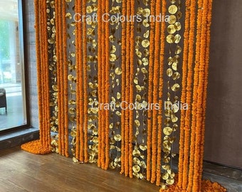 May include: A decorative backdrop made of orange marigold flower garlands and gold paper circles. The garlands are strung together and hang from a ceiling, creating a curtain-like effect. The backdrop is perfect for a wedding or other special event.
