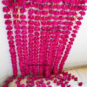 May include: Rows of vibrant fuchsia artificial marigold flower garlands. The garlands are strung together with green stems and gold wire, creating a decorative floral display. The text "Craft colours India" is visible on the image.