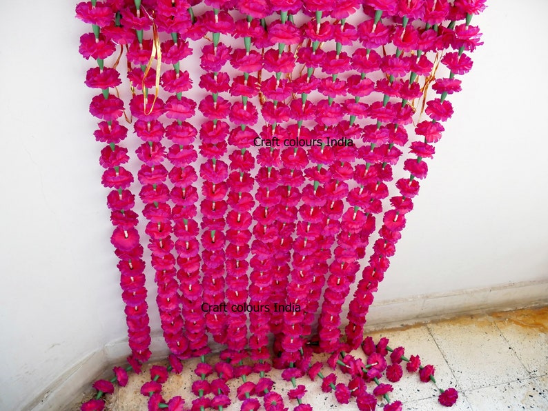 May include: Rows of vibrant fuchsia artificial marigold flower garlands. The garlands are strung together with green stems and gold wire, creating a decorative floral display. The text "Craft colours India" is visible on the image.