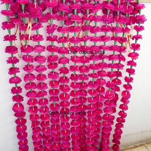 May include: A vibrant magenta floral garland, composed of numerous flower strands. Each strand features closely spaced, round magenta flowers with green stems. The garland is suspended, creating a decorative, eye-catching display. The text "Craft colours India" is visible.