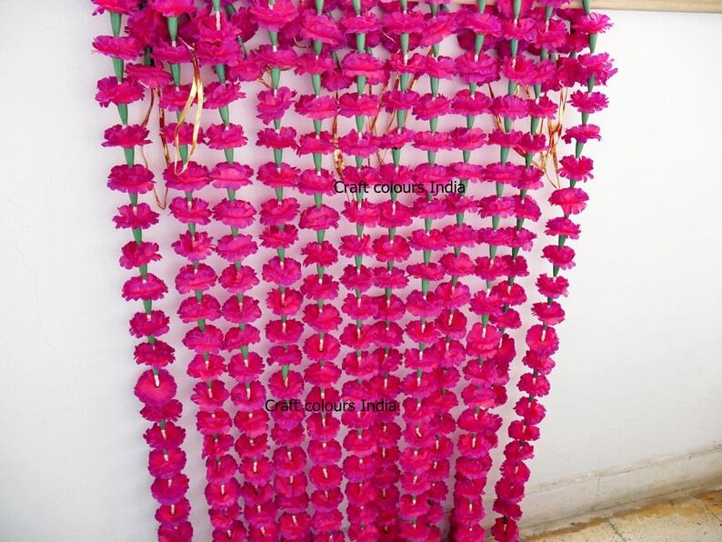 May include: A vibrant magenta floral garland, composed of numerous flower strands. Each strand features closely spaced, round magenta flowers with green stems. The garland is suspended, creating a decorative, eye-catching display. The text "Craft colours India" is visible.