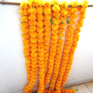 May include: A garland of bright yellow marigold flowers, strung together in a long strand. The garland is hanging on a white wall.