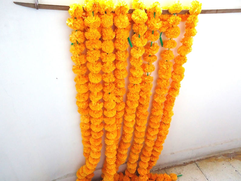 May include: A garland of bright yellow marigold flowers, strung together in a long strand. The garland is hanging on a white wall.