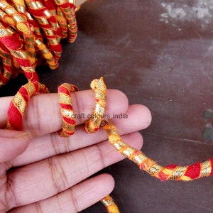 May include: Red and gold braided trim with a metallic sheen. The trim is twisted and looped in a hand.