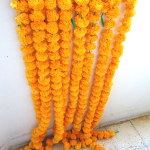 May include: A string of yellow marigold flower garlands hanging on a white wall. The garlands are made of paper and are strung together with green string.