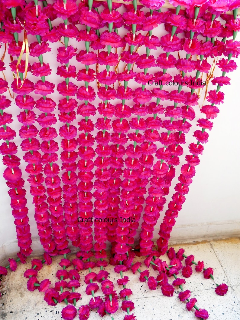 May include: Rows of vibrant fuchsia artificial marigold flower garlands hang from a wooden beam. The garlands are suspended in vertical lines, with the text "Craft colours India" visible on the right side.