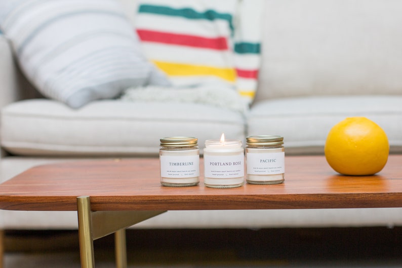May include: Three glass candle jars with gold lids on a wooden table. The candles are labeled "Timberline", "Portland Rose", and "Pacific".