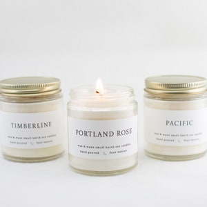 May include: Three glass candle jars with gold lids. The candles are labeled "Timberline", "Portland Rose", and "Pacific". Each jar has a white label with black text that reads "wax & wane small batch soy candles, hand poured, four ounces". The candle in the center is lit.