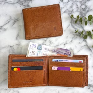May include: Two brown leather wallets, one open and one closed, on a marble surface. The open wallet holds banknotes, credit cards, and a UK driving licence. The closed wallet features a small embossed design.