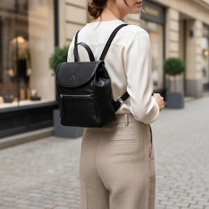 May include: A black leather backpack with a flap, top handle, and adjustable straps. It has a gold-coloured zip and an embossed emblem on the flap. The bag is worn by a person in a white blouse and beige trousers, walking outdoors.