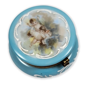 May include: A round, light blue trinket box with a hinged lid. The lid features a painted scene of cherubs in a cloudy sky, framed by a white, ornate border. The box has a gold-colored clasp and white decorative accents.