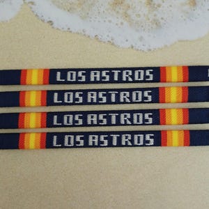 May include: Four navy blue woven bracelets with white text that reads "LOS ASTROS" and orange and yellow stripes.
