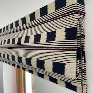 Könnte beinhalten: Ein marineblauer und cremefarbener römischer Rollo mit einem geometrischen Muster aus Quadraten und horizontalen Streifen. Ein dünner roter Streifen akzentuiert die Oberkante. Der Rollo ist an einem Fensterrahmen montiert.