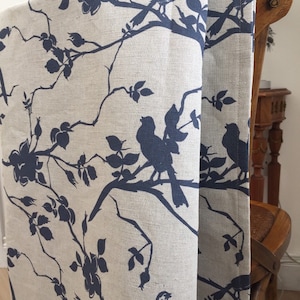 May include: A linen tablecloth with a blue and white floral pattern featuring silhouettes of birds on branches. The tablecloth is draped over a wooden chair.