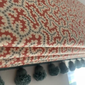 May include: Close-up of a Roman shade with a floral pattern in red, cream, and gray. The shade is trimmed with teal pom-poms along the bottom edge. The fabric is folded in horizontal pleats.