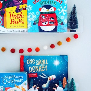 May include: A white wall with a garland of colorful felt balls. There are three bookshelves with children's books on them. The books are titled "Jingle Bugs", "Ugly Sweater", and "One Small Donkey". There are two small artificial Christmas trees on the shelves.